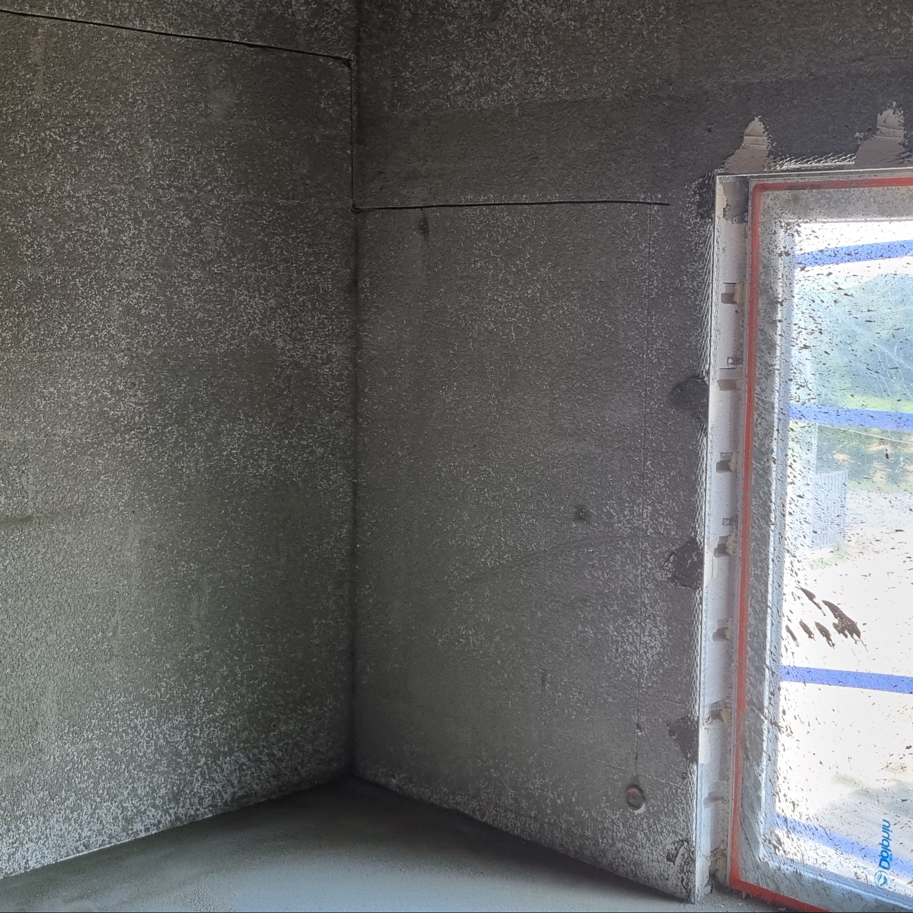 Wnętrze w trakcie prac wykończeniowych: ściany pokryte świeżo nałożonym szarym tynkiem, widoczne zarysowania i nierówności faktury, okno zabezpieczone folią z zabrudzeniami.