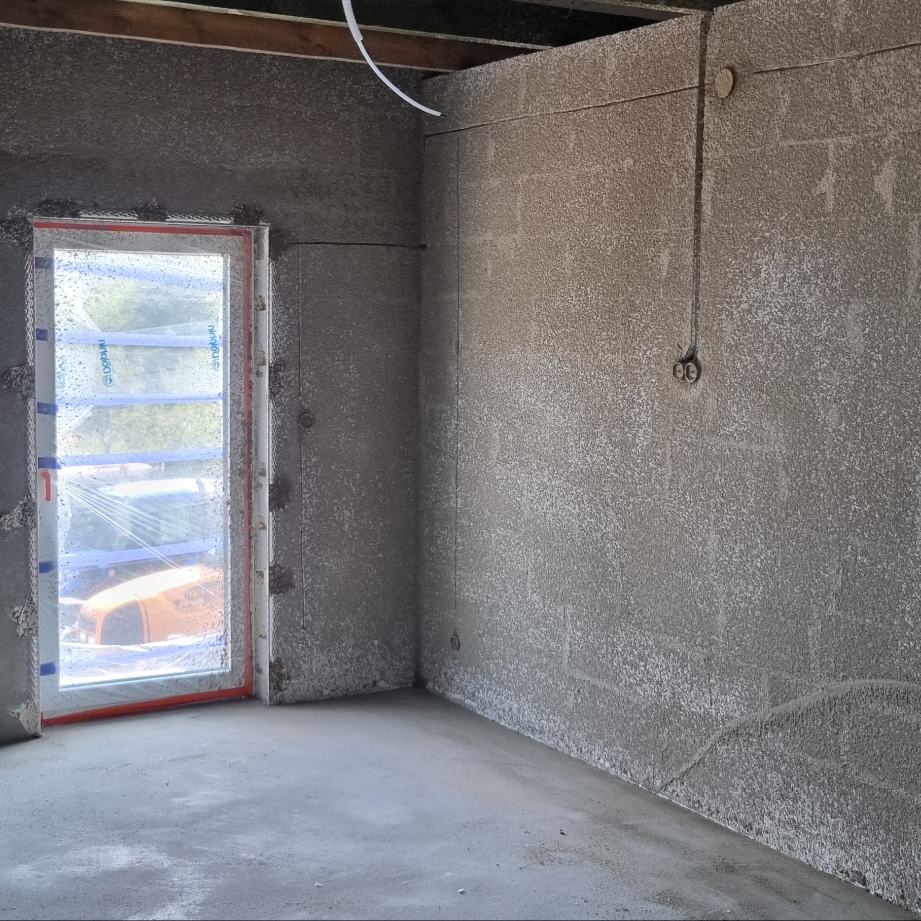 Wnętrze pomieszczenia po nałożeniu szarego tynku maszynowego na ściany i podłogę, widoczne zabezpieczone okno z folią ochronną.