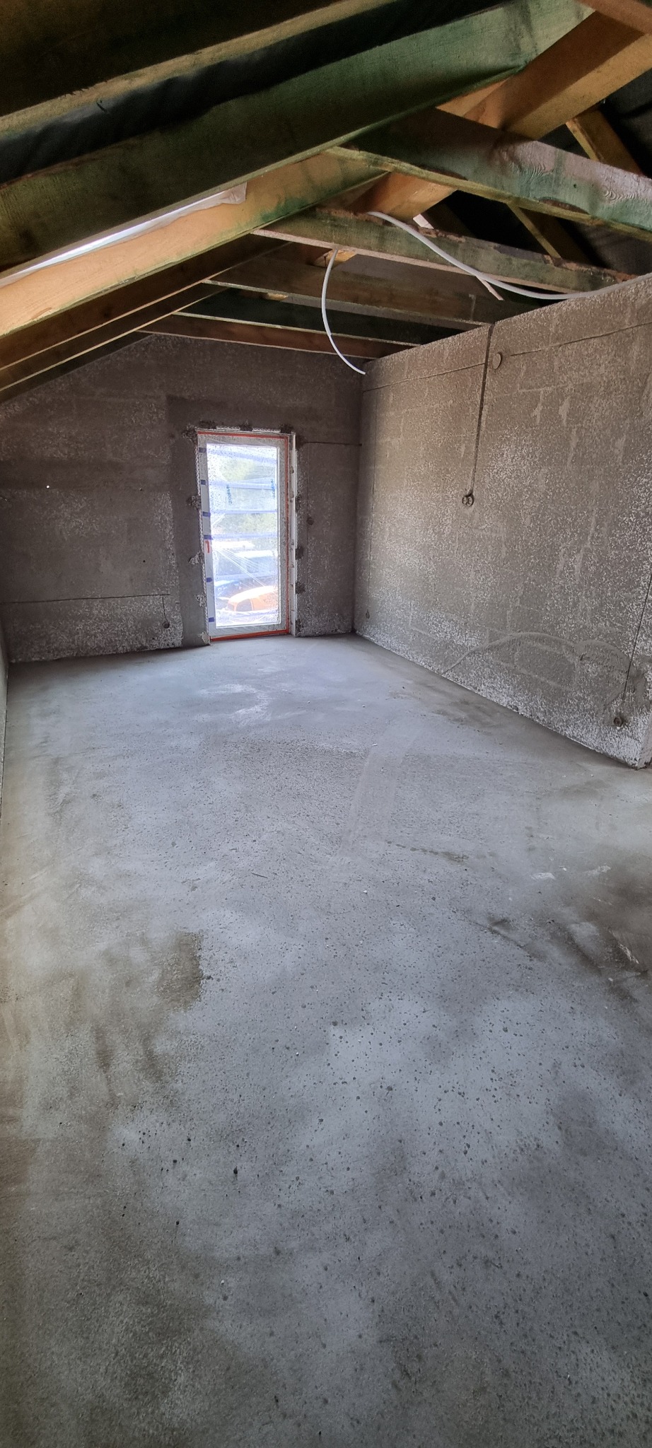 Surowe wnętrze poddasza ze świeżo nałożonymi szarymi tynkami na ścianach i podłodze, widoczne drewniane elementy konstrukcji dachu oraz okno zabezpieczone folią.