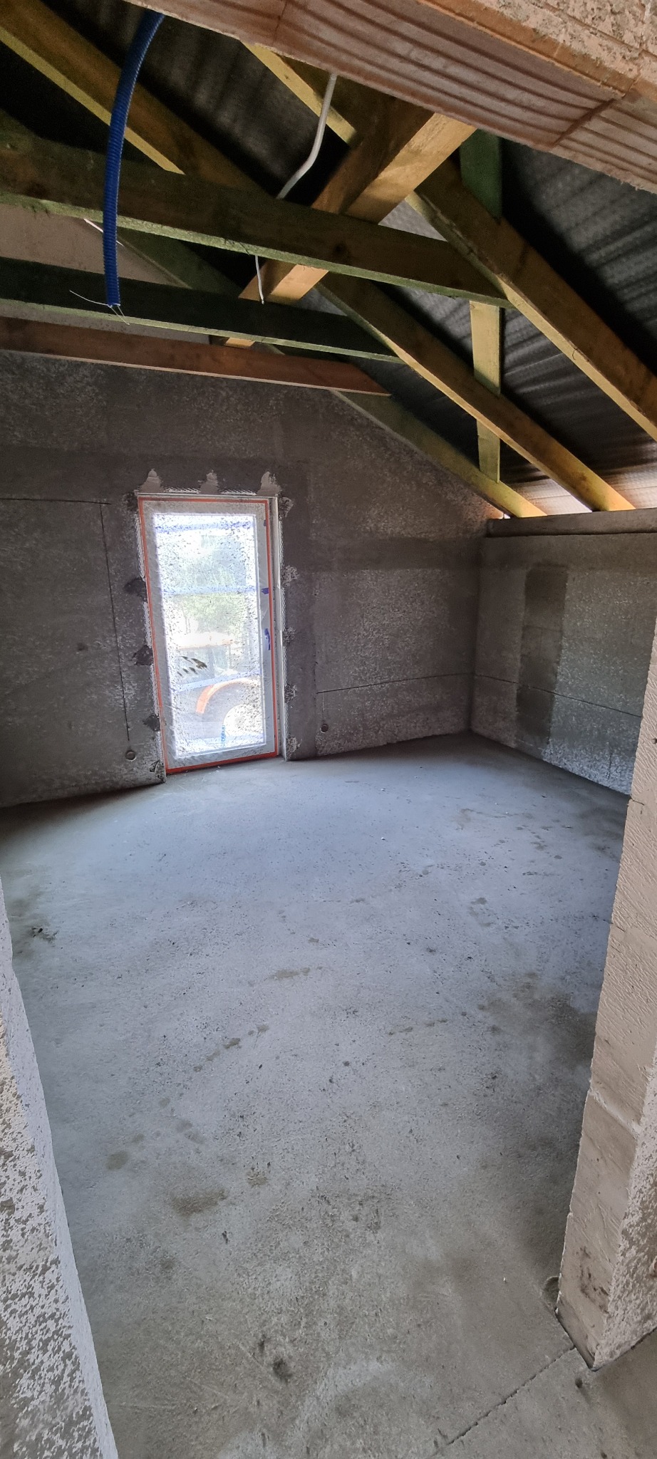 Surowe wnętrze poddasza po nałożeniu tynku maszynowego, widoczne drewniane krokwie dachu, okno z pomarańczową ramką i niebieski przewód instalacyjny pod sufitem.