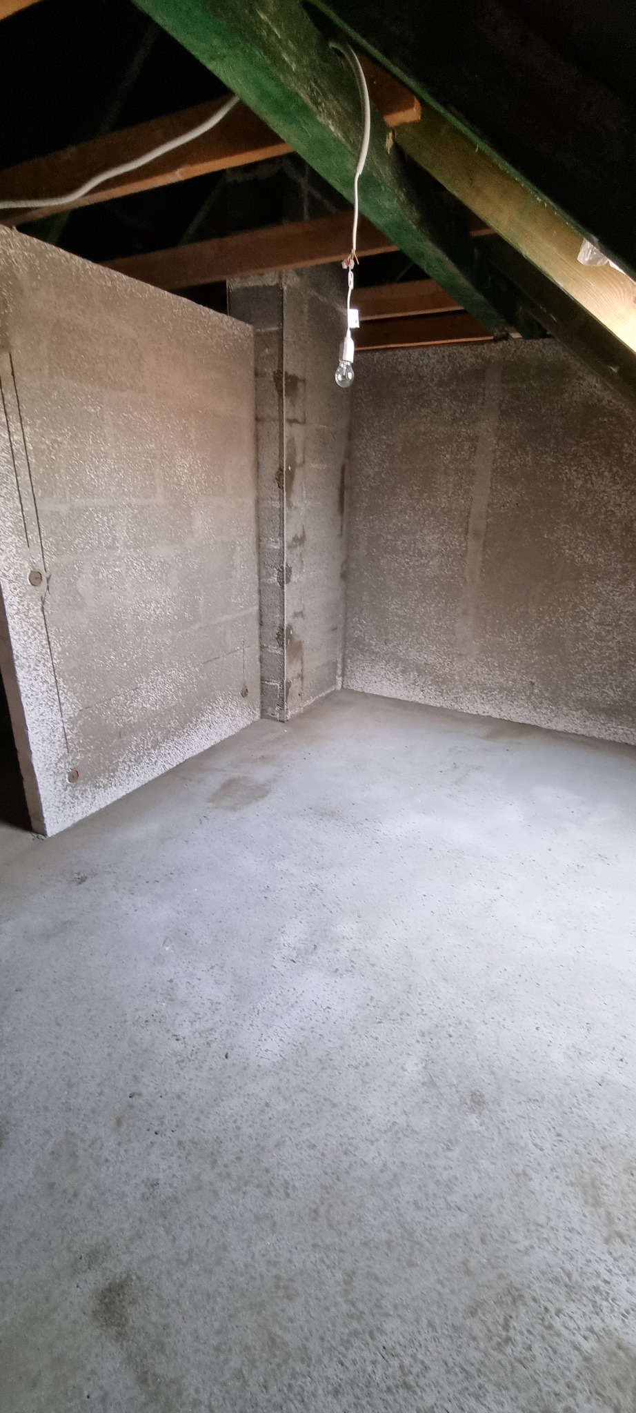 Pokój na poddaszu po tynkowaniu maszynowym ścian i wylewce podłogi, widoczne surowe drewniane belki stropowe i prowizoryczne oświetlenie.