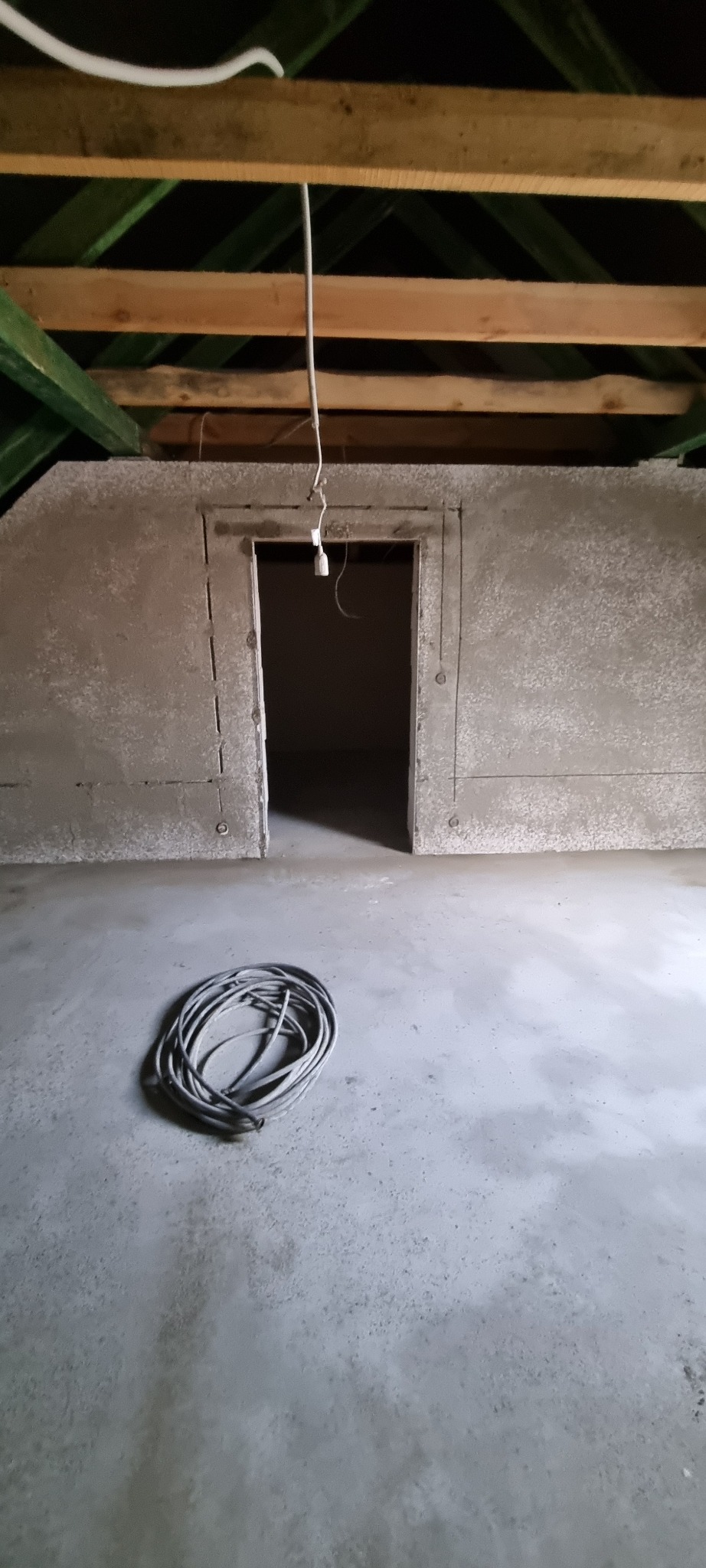 Wnętrze poddasza po nałożeniu tynku maszynowego, widoczne drewniane elementy konstrukcji dachu, przygotowany otwór drzwiowy i zwój kabla na posadzce.