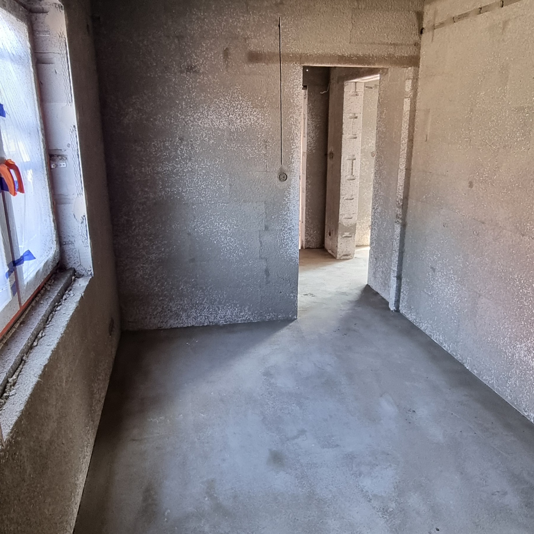 Wnętrze w stanie deweloperskim z nałożonymi szarymi tynkami na ścianach i wylewką na podłodze, widoczne zabezpieczone okno folią i przejście do kolejnych pomieszczeń.