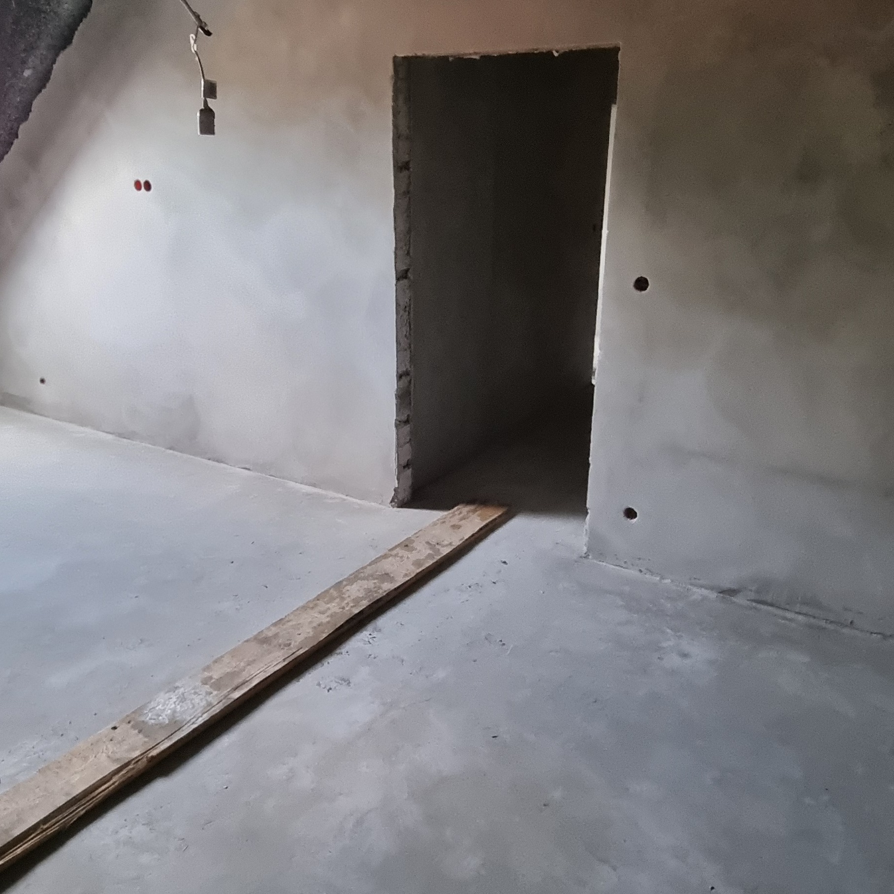 Surowe wnętrze po nałożeniu tynku maszynowego; widoczna drewniana listwa na szarej, gładkiej podłodze i otwór drzwiowy w ścianie.