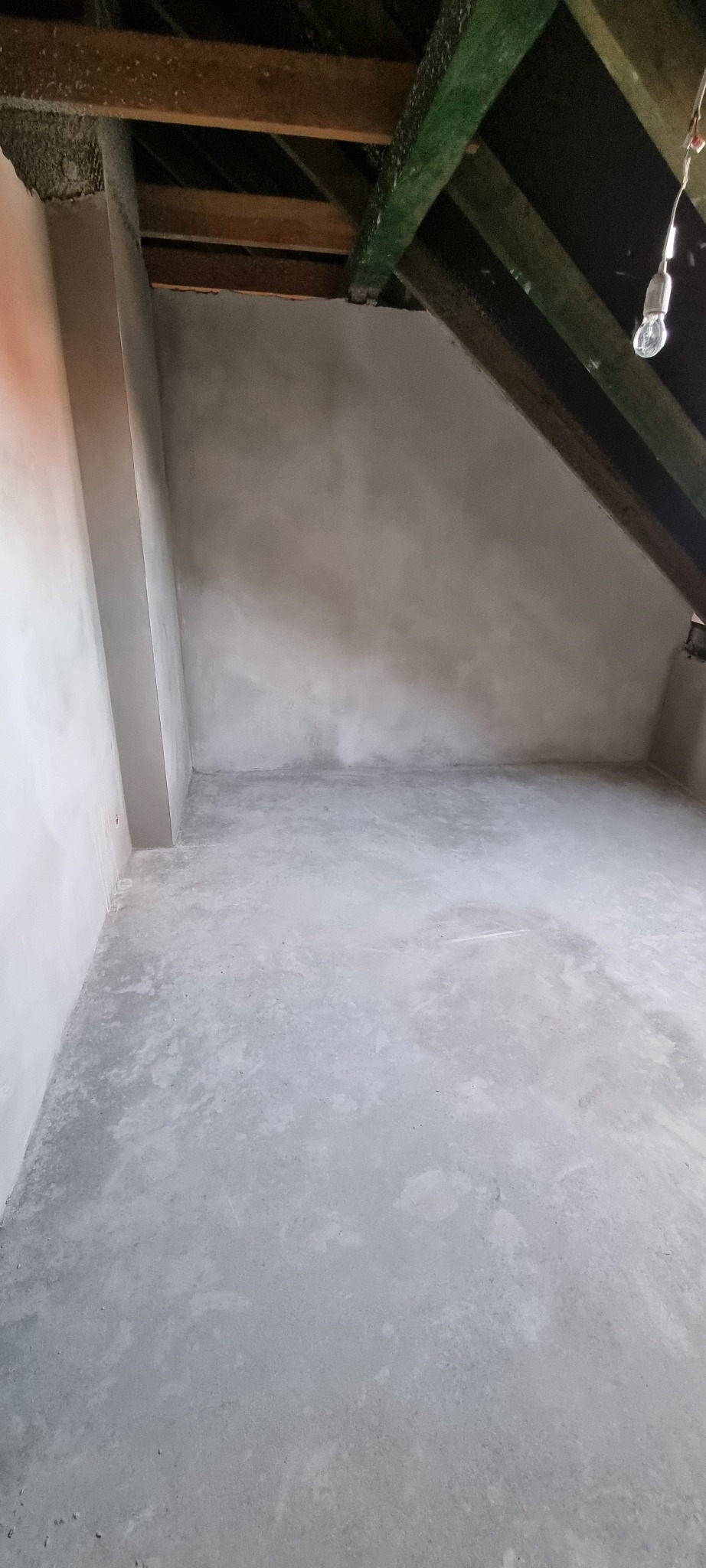 Wnętrze poddasza po nałożeniu gładzi cementowej na ściany i podłogę, widoczne drewniane elementy konstrukcyjne dachu i wisząca żarówka.
