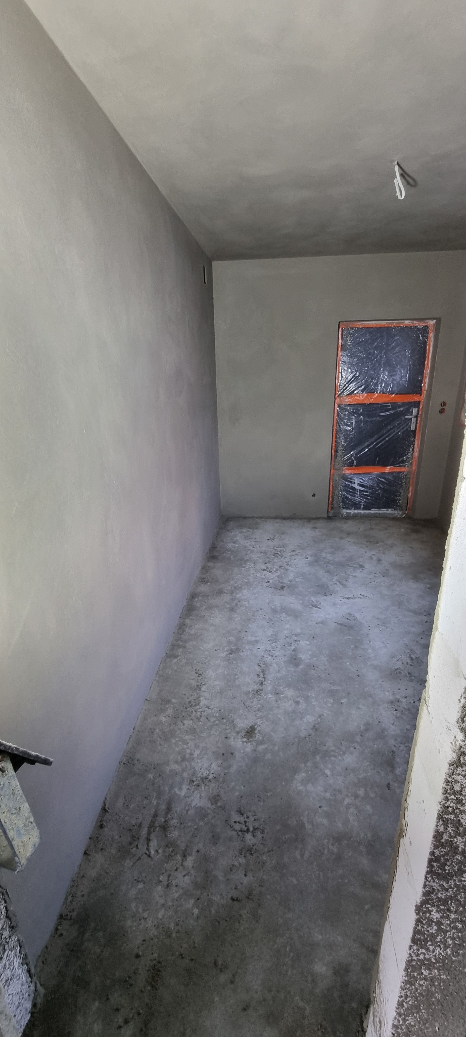 Wnętrze pomieszczenia po nałożeniu świeżych, szarych tynków na ścianach i suficie, drzwi zabezpieczone folią ochronną, surowy beton na podłodze.