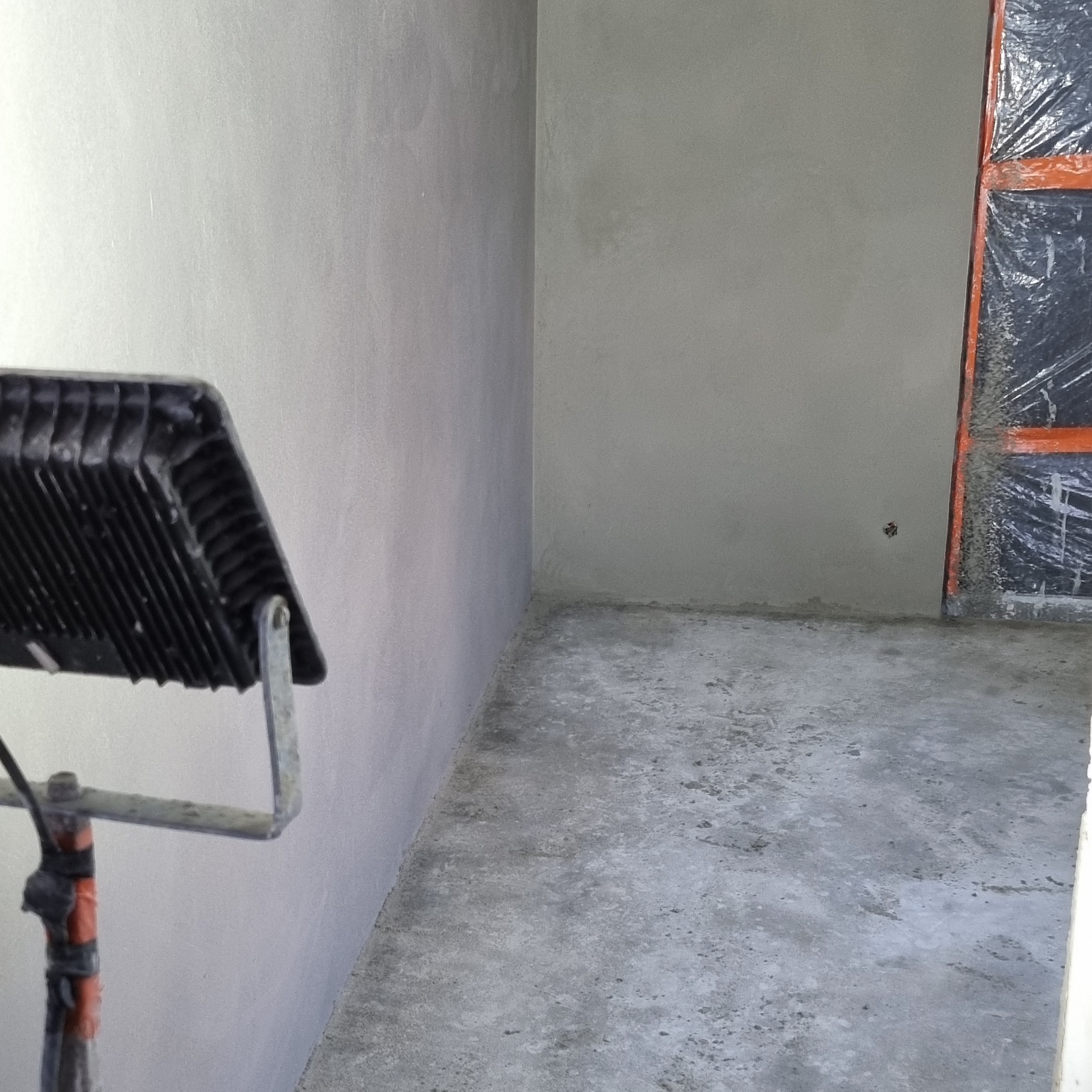 Świeżo nałożony, jasnoszary tynk na ścianach w rogu pomieszczenia, oświetlony lampą budowlaną. Okno zabezpieczone folią w tle.