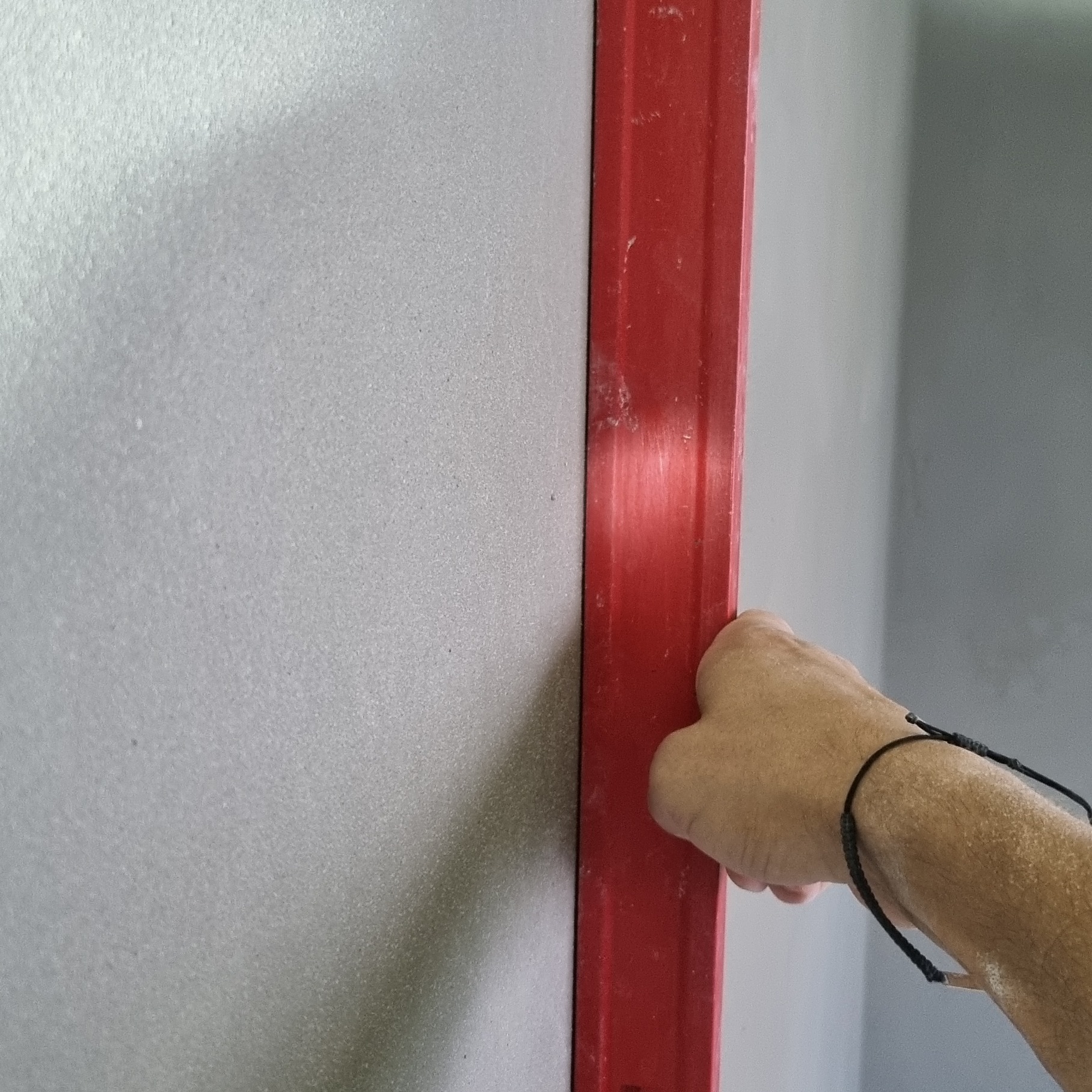 Czerwona poziomica przykładana do świeżo nałożonego, szarego tynku maszynowego na ścianie, widoczna dłoń z czarną bransoletką.