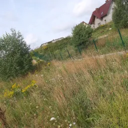 Zarośnięty teren z zielonym ogrodzeniem siatkowym, w tle dom z czerwoną dachówką na wzniesieniu.