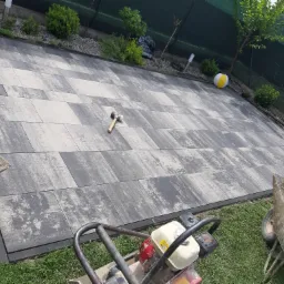 Ułożona nawierzchnia z szarej i białej kostki brukowej, widoczny ubijak wibracyjny, taczka z piaskiem, gumowy młotek, w tle trawnik i ogrodzenie.