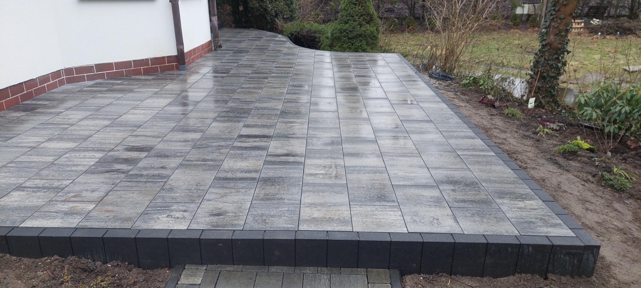 Szeroki taras z szarej kostki brukowej, obrzeża wykończone czarną kostką, widok na ogród z krzewami i drzewami, po deszczu.