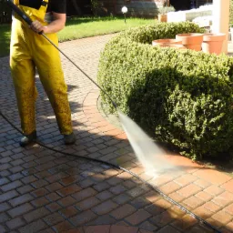 Osoba w żółtym kombinezonie roboczym czyści kostkę brukową na podwórku za pomocą myjki ciśnieniowej, w tle żywopłot i donice.