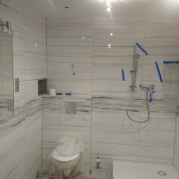 Konkretna Ekipa Remontowa - Łazienka w trakcie remontu, widoczne białe płytki imitujące marmur, zabezpieczona toaleta folią, prysznic z niebieską taśmą malarską.