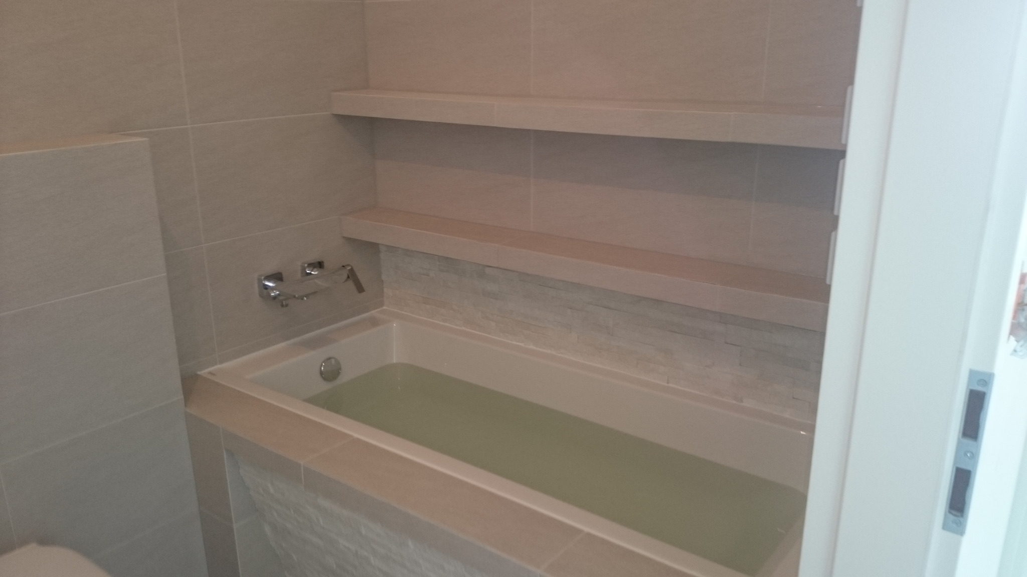 Wykończona łazienka z wanną wypełnioną wodą, jasne płytki na ścianach, dwie półki nad wanną i dekoracyjny panel z kamienia. Widoczny fragment baterii wannowej.