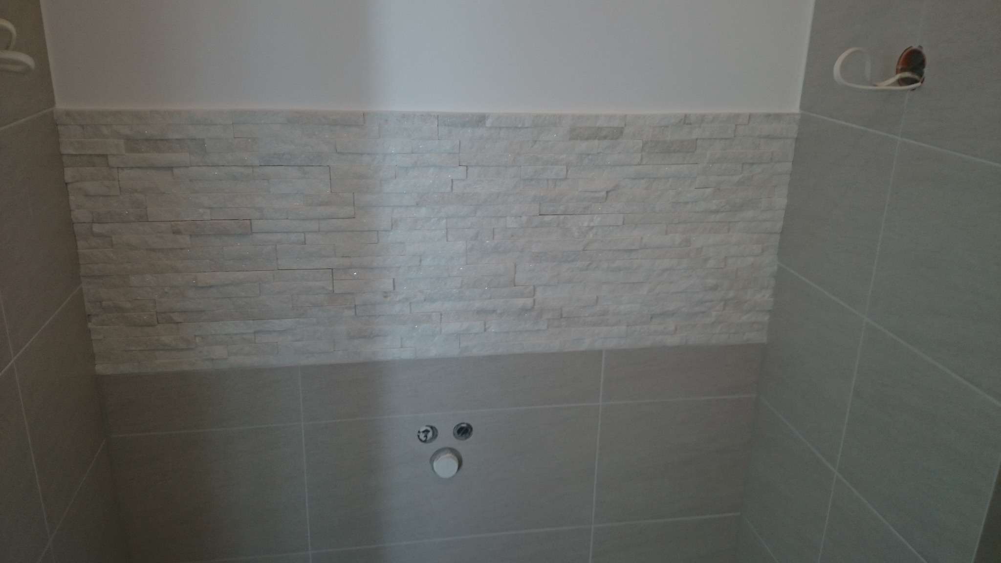 Fragment ściany w łazience z dekoracyjną, białą okładziną imitującą kamień powyżej szarej, gładkiej płytki; widoczne przyłącza kanalizacyjne i wystające kable elektryczne.