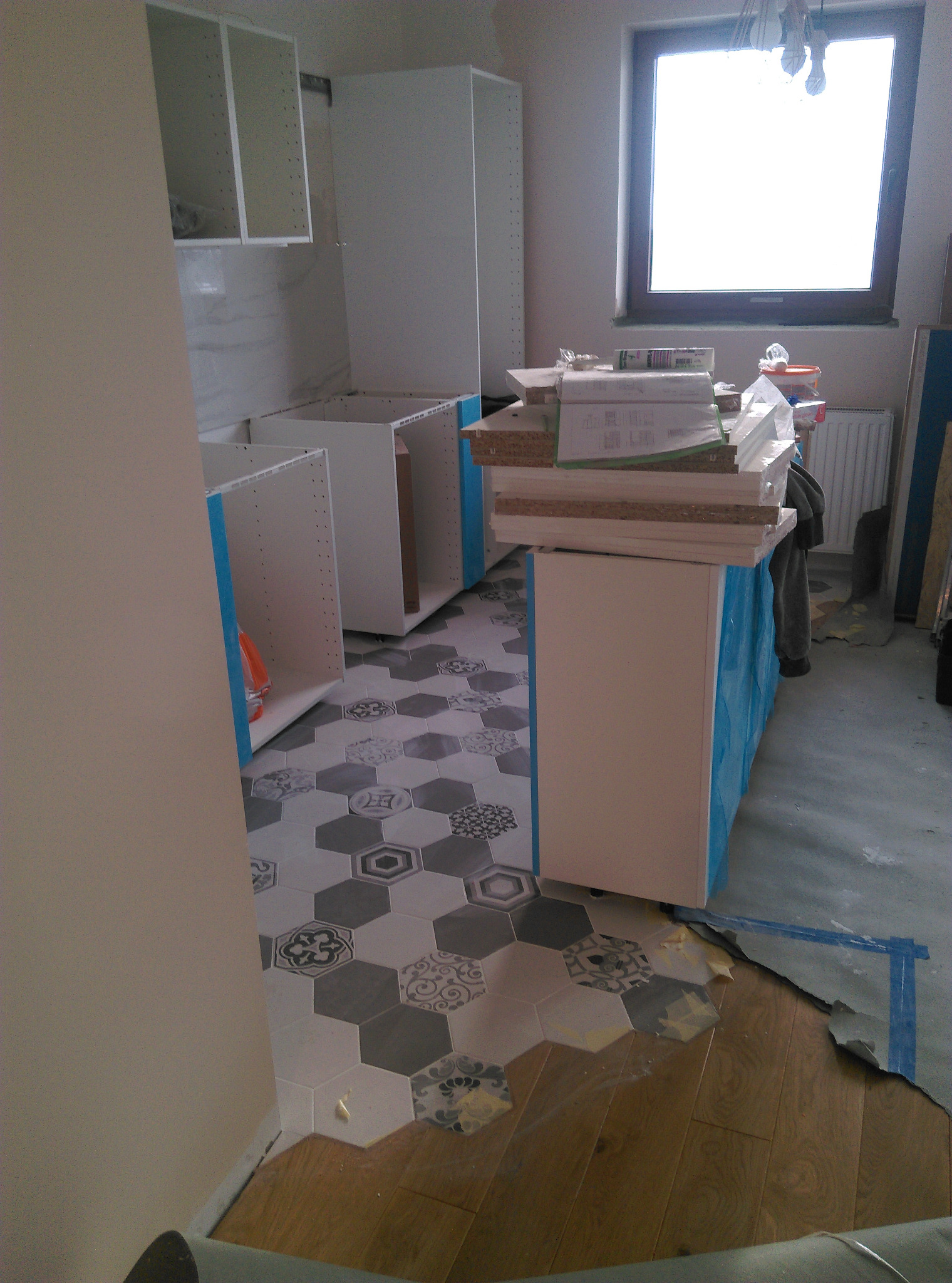 Montaż szafek kuchennych z zabezpieczeniem folią, widoczne płytki heksagonalne na podłodze i fragment parkietu, etap prac wykończeniowych.