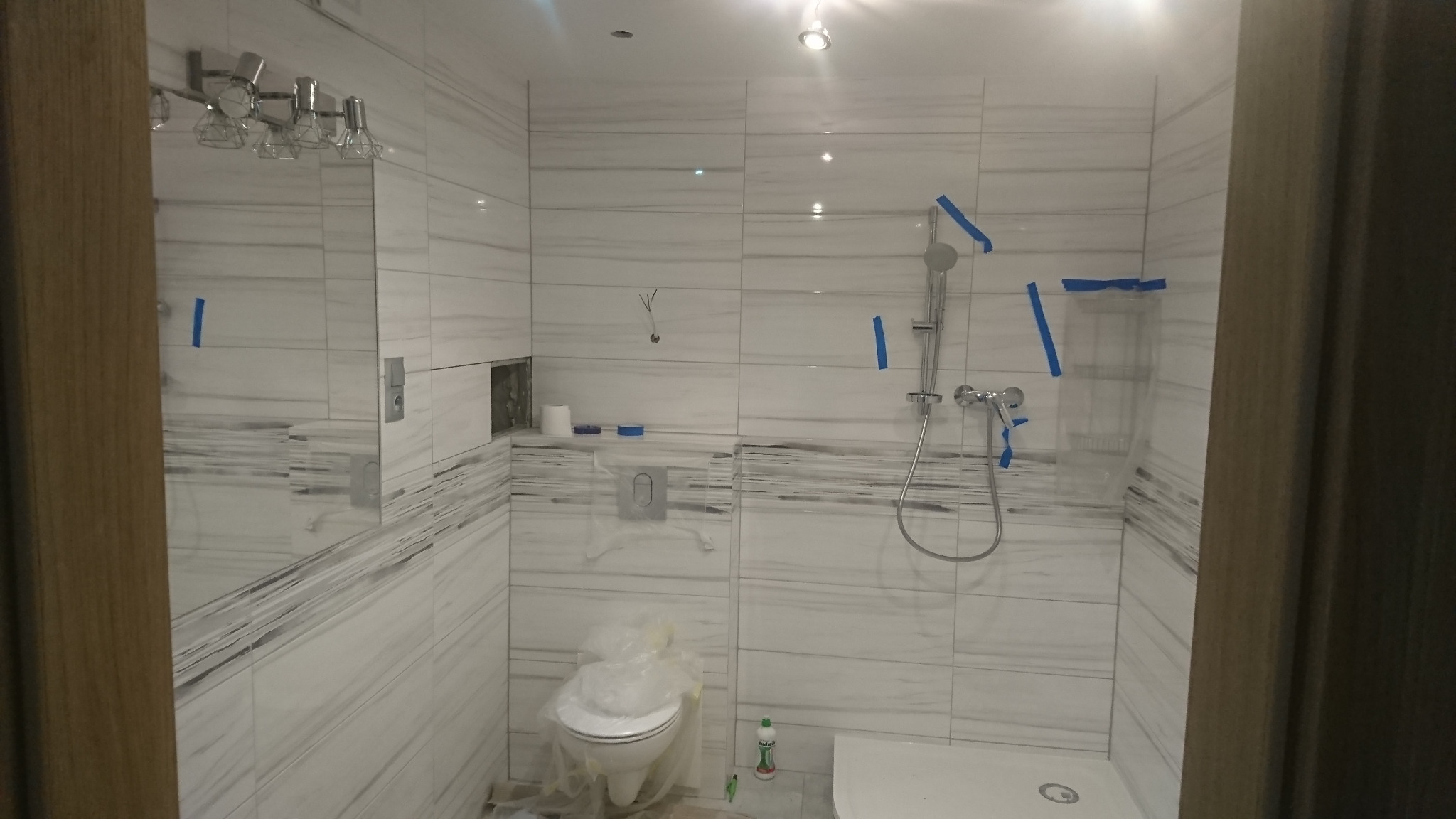 Łazienka w trakcie remontu, widoczne białe płytki imitujące marmur, zabezpieczona toaleta folią, prysznic z niebieską taśmą malarską.