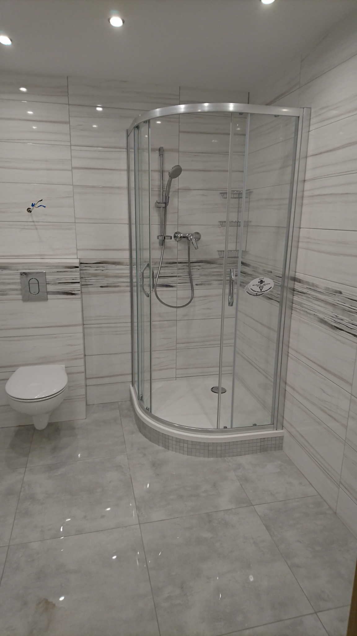 Nowoczesna łazienka wykończona w szaro-białej kolorystyce, z kabiną prysznicową z zaokrąglonymi drzwiami, wiszącą toaletą i płytkami imitującymi marmur na ścianach, oraz dużymi płytkami podłogowymi.