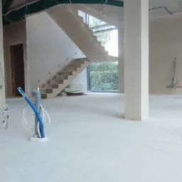 Wnętrze w stanie surowym: gładka, jasna wylewka podłogowa, kolumna nośna, betonowe schody z oknem na ogród, instalacje elektryczne i hydrauliczne wystające z podłogi.