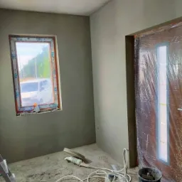 Wnętrze pokoju w trakcie prac wykończeniowych: ściany pokryte świeżym tynkiem, okno z widokiem na zewnątrz, drzwi zabezpieczone folią, na podłodze leżą narzędzia i przewody, widoczna drabina oparta...