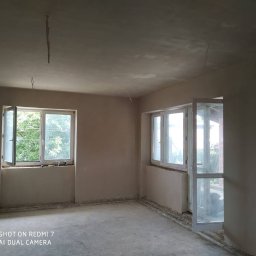 Boks.o.o.s Mikołaj - Pokój w trakcie remontu: świeżo położona warstwa gładzi na ścianach i suficie, otwarte okno z widokiem na zieleń. Widoczne niedociągnięcia i kable wystające z sufitu.