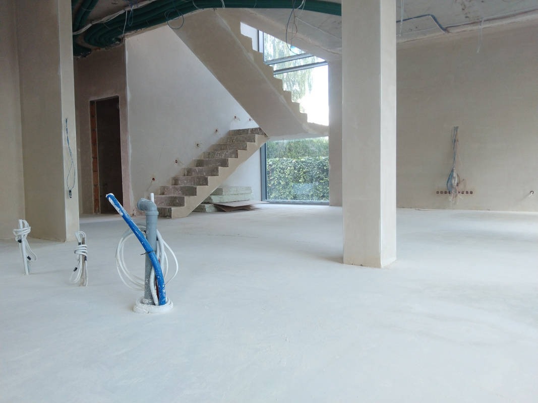 Wnętrze w stanie surowym: gładka, jasna wylewka podłogowa, kolumna nośna, betonowe schody z oknem na ogród, instalacje elektryczne i hydrauliczne wystające z podłogi.
