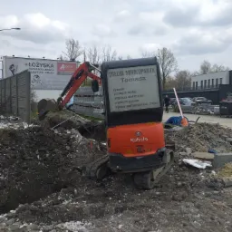 Pomarańczowa minikoparka Kubota w trakcie prac ziemnych, widoczny wykop w ziemi i reklama usług minikoparką z numerem telefonu.