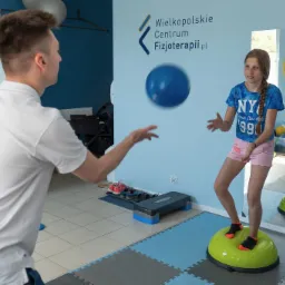 Młoda dziewczyna ćwiczy równowagę na zielonej platformie w gabinecie, podczas gdy fizjoterapeuta rzuca do niej niebieską piłką. W tle logo Wielkopolskiego Centrum Fizjoterapii i lustro z kolorowym...