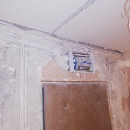Vitalii Kurtash - Rozprowadzenie instalacji elektrycznej w trakcie remontu, widoczne kable w peszlach na ścianie i suficie, otwarta skrzynka bezpiecznikowa z podłączonymi przewodami, surowe ściany przed wykończeniem.