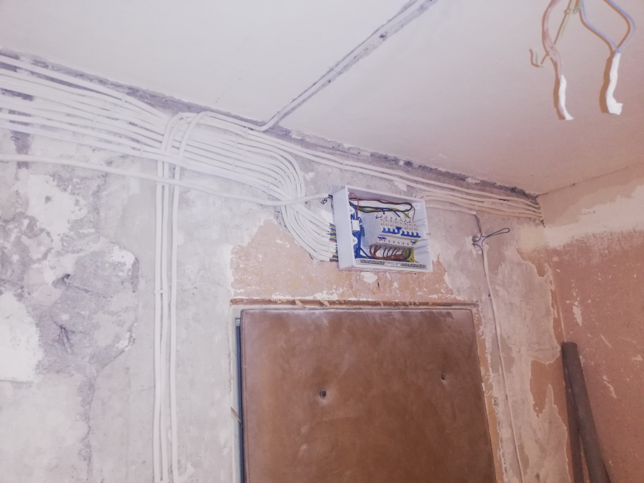 Rozprowadzenie instalacji elektrycznej w trakcie remontu, widoczne kable w peszlach na ścianie i suficie, otwarta skrzynka bezpiecznikowa z podłączonymi przewodami, surowe ściany przed wykończeniem.