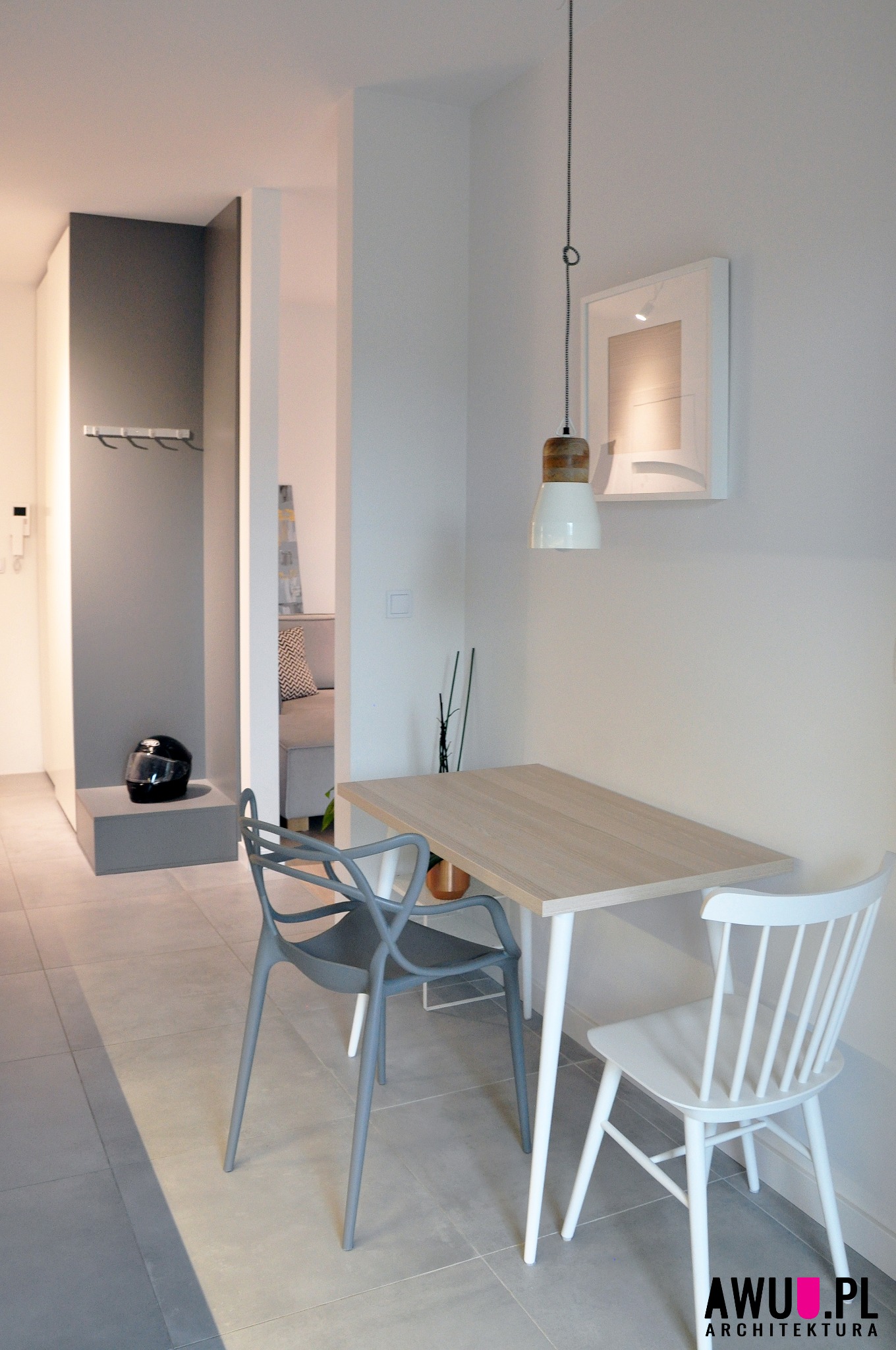 Jasna jadalnia z drewnianym stołem, krzesłem w stylu Windsor i szarym krzesłem designerskim. W tle widoczny przedpokój z czarnym kaskiem motocyklowym i wieszakami.