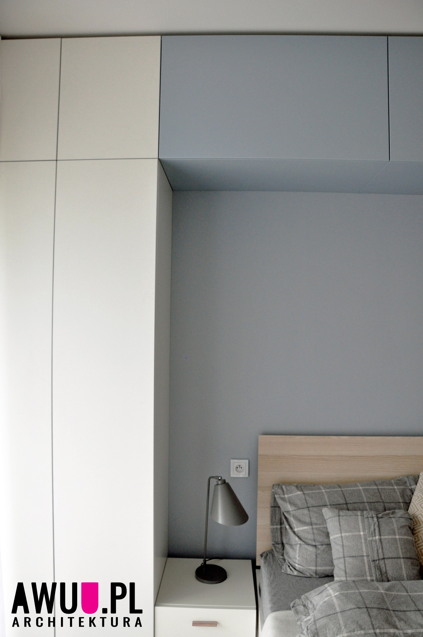 Minimalistyczna sypialnia z szarą ścianą, drewnianym zagłówkiem, pościelą w kratę, szafką nocną z lampką i zabudową szafy z białymi i szarymi frontami.