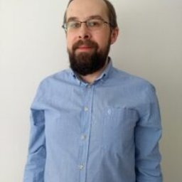 Pisanie i Marketing - Mężczyzna w okularach i błękitnej koszuli, z brodą, stojący na jasnym tle.