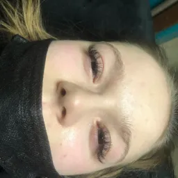 Zbliżenie na twarz kobiety z przedłużonymi rzęsami, widoczne świeżo wykonane, gęste rzęsy, dolna część twarzy zasłonięta czarną maską ochronną, włosy blond.