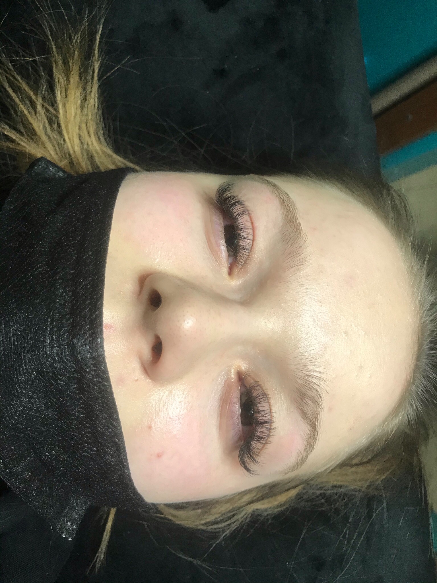 Zbliżenie na twarz kobiety z przedłużonymi rzęsami, widoczne świeżo wykonane, gęste rzęsy, dolna część twarzy zasłonięta czarną maską ochronną, włosy blond.