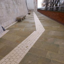 Firma Kames - Kamienny chodnik z jasnym pasem kostki, prowadzący wzdłuż muru i ceglanego ogrodzenia, z kamiennymi ławkami w tle.