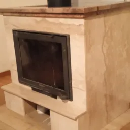 Obudowa kominkowa z beżowego kamienia z widoczną czarną wkładką kominkową, drewnianą podłogą i fragmentem mebla w tle.