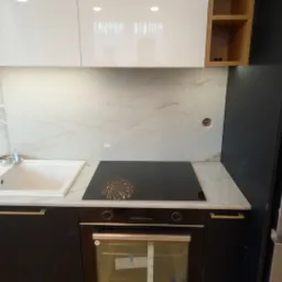 Nowoczesna kuchnia z czarnymi szafkami, złotymi uchwytami, białym blatem z marmurowym wzorem, płytą indukcyjną i piekarnikiem. Nad blatem białe szafki wiszące i marmurowa ściana. Widoczny fragment...