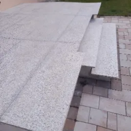 Granitowe schody wejściowe z tarasem, wyłożone jasnoszarą kostką, widok z boku na tle trawnika i elewacji budynku.