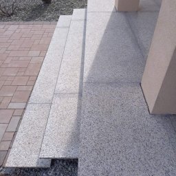 Firma Kames - Zewnętrzne schody z jasnoszarego granitu prowadzące do wejścia, z widocznymi słupami podtrzymującymi zadaszenie, obok ścieżka z czerwonej kostki brukowej i ozdobny żwir.