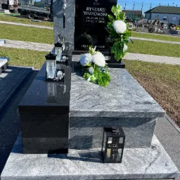 Granitowy nagrobek z czarną tablicą, białymi kwiatami i lampionami na cmentarzu. Widoczne nagrobki w tle, jasne oświetlenie słoneczne.