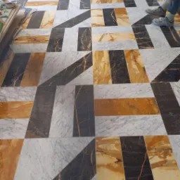 Wzorzysta podłoga z ułożonych elementów kamiennych w odcieniach bieli, brązu i czerni, tworząca geometryczny design z widocznymi fragmentami nóg osoby stojącej na niej.