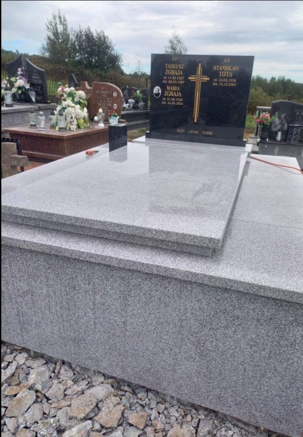 Jasnoszary granitowy nagrobek z czarną tablicą z wygrawerowanym złotym krzyżem i napisami, znajdujący się na cmentarzu, w tle inne groby i drzewa.