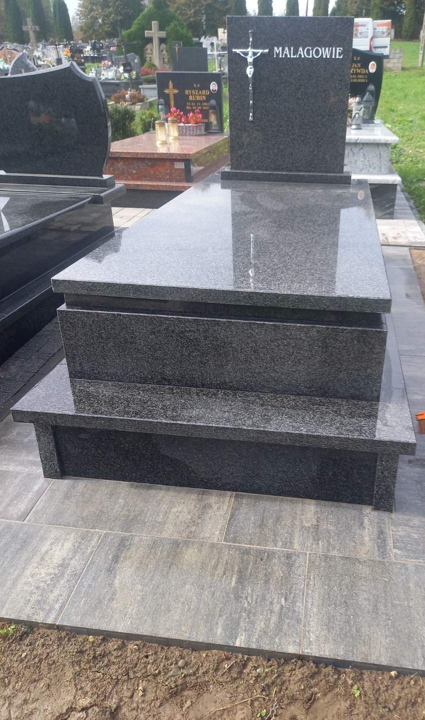 Nowoczesny, trzyczęściowy nagrobek z ciemnego granitu, z inskrypcją 'Malagowie' i metalowym krucyfiksem, otoczony innymi nagrobkami na cmentarzu.