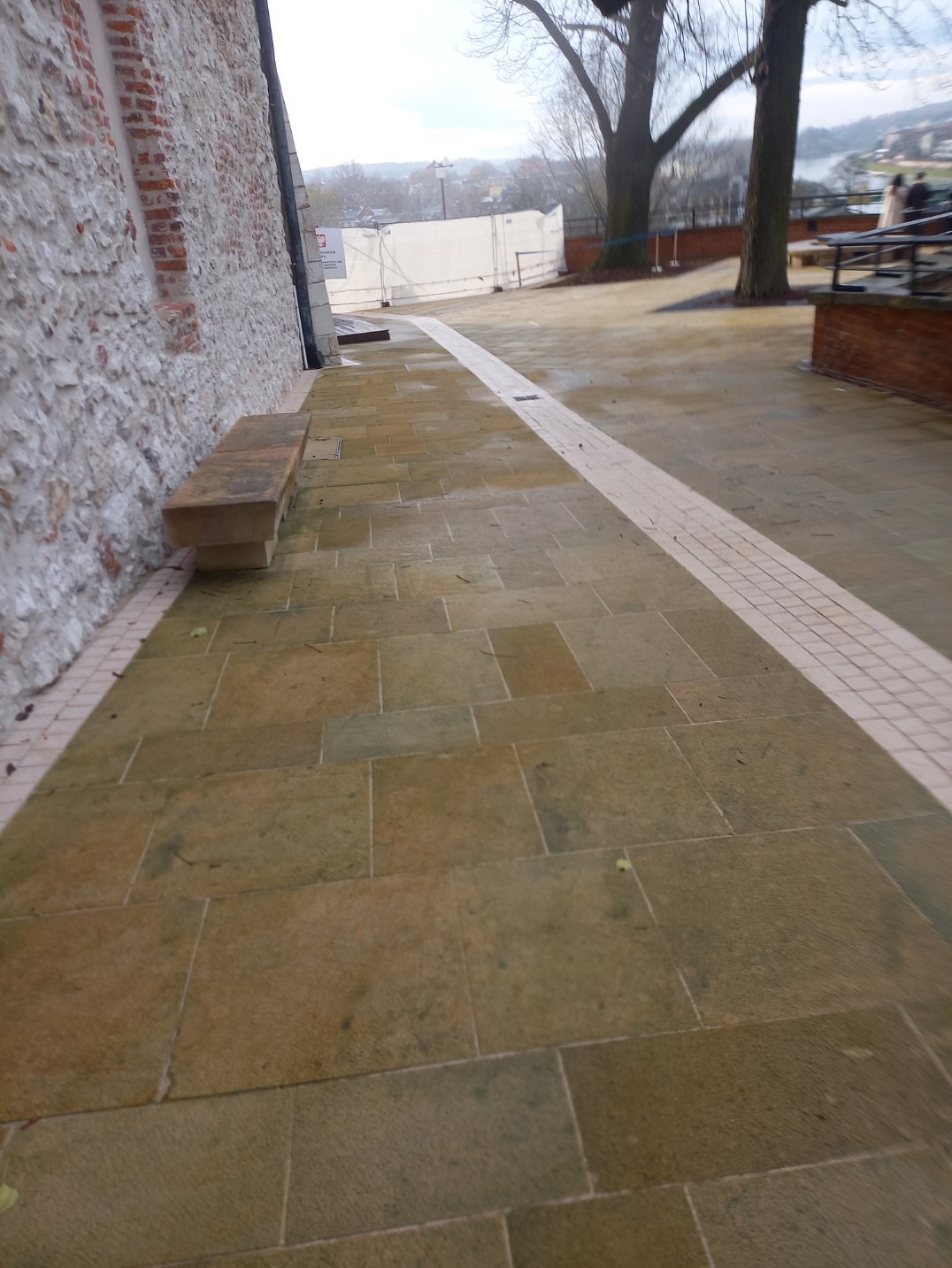 Kamienny chodnik z ławką przylegający do kamiennej ściany budynku, z widoczną linią z kostki brukowej i drzewami w tle.