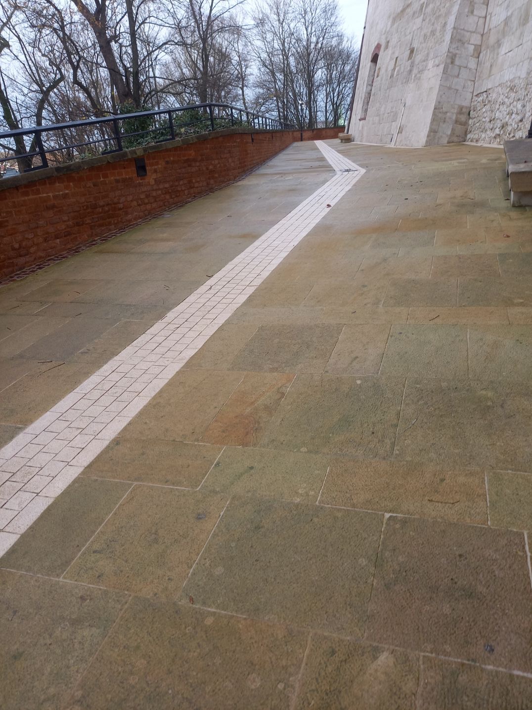 Kamienny taras prowadzący wzdłuż ceglanego murku i kamiennej ściany budynku, z wyraźnie zaznaczoną ścieżką z jasnej kostki, ułożony na wzniesieniu.