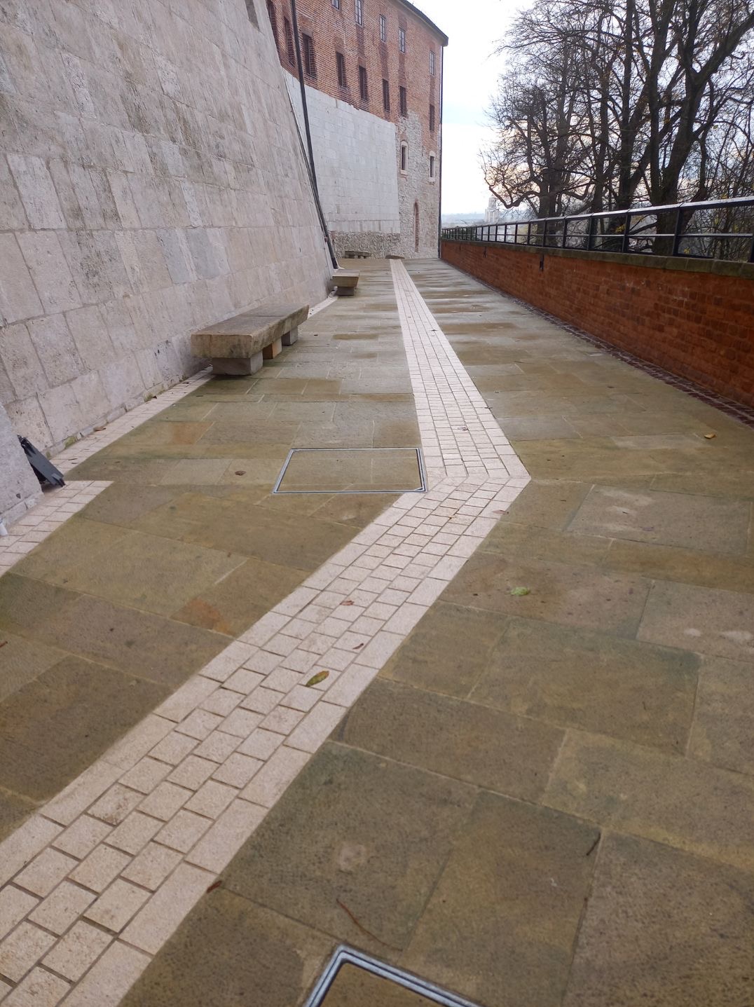Kamienny chodnik z jasnym pasem kostki, prowadzący wzdłuż muru i ceglanego ogrodzenia, z kamiennymi ławkami w tle.