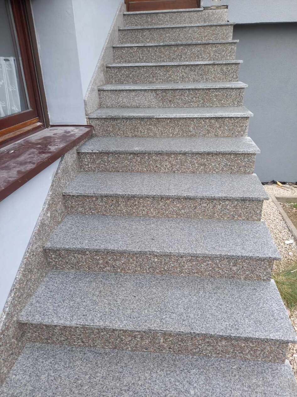 Zewnętrzne schody z szarego kamienia ziarnistego, prowadzące od okna z brązową ramą ku górze, z widocznymi drobnymi kamyczkami obok.