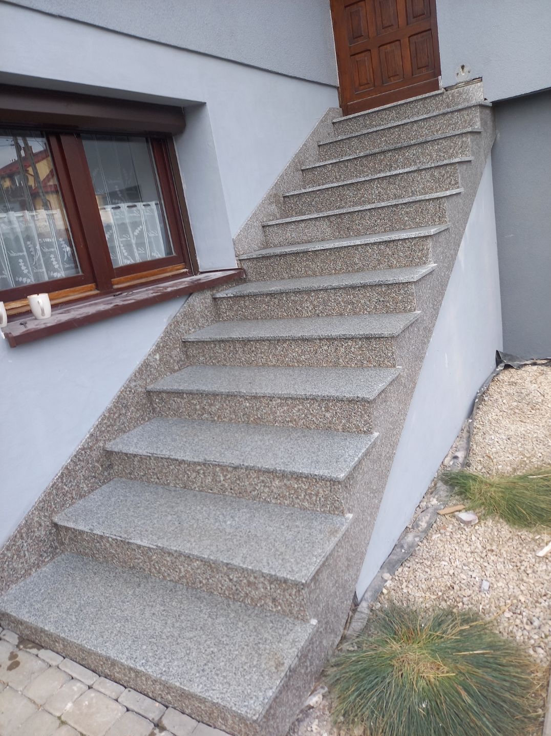 Zewnętrzne schody z szarego kamienia o drobnej fakturze, prowadzące do drewnianych drzwi wejściowych, z widocznym oknem z brązową ramą i drobnymi dekoracjami na parapecie.