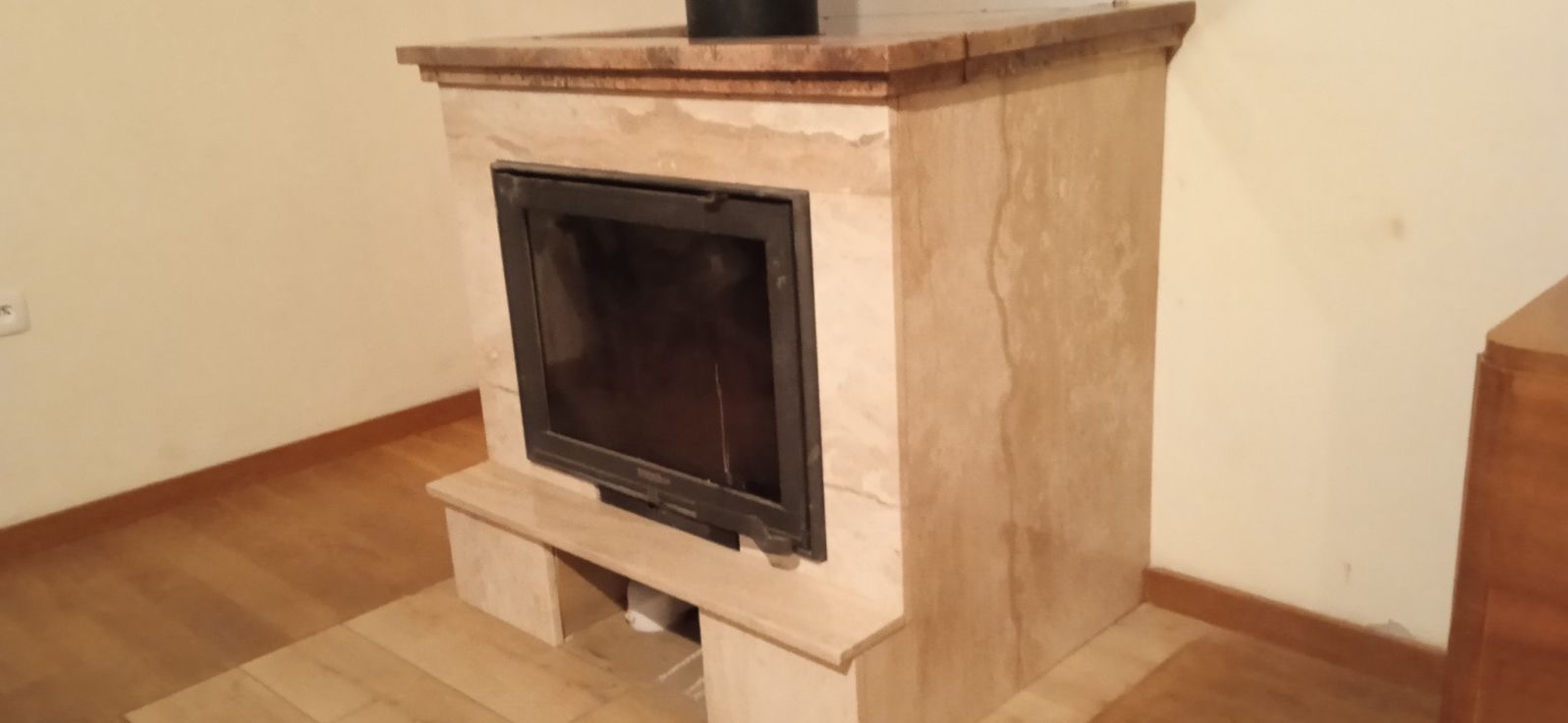 Obudowa kominkowa z beżowego kamienia z widoczną czarną wkładką kominkową, drewnianą podłogą i fragmentem mebla w tle.
